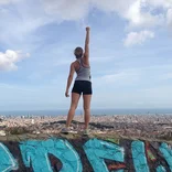 student standing on top of a graffitied ledge, punching the sky like Superman with the Barcelona skyline in the distance