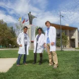 three students wearing lab coats standing on a grassy lawn in front of a building