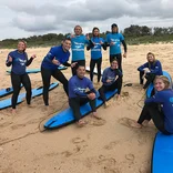 group of students wearing wetsuits sitting on and standing near surf boards on the beach