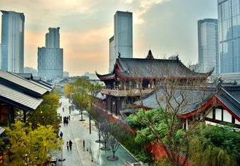 An image of Chengdu is shown. In the background, large city buildings can be seen, and in the foreground there are traditional Chinese buildings. On the left side of the image is a street lines with trees with people walking along it.