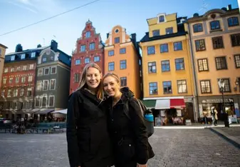 Two female students are standing in an old town square with colorful historical buildings behind them.