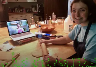 Image of a student with a rolling pin. A laptop in the background shows another student's face. 