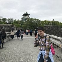 The image is a vertical picture of a girl standing on a bridge, making a peace sign with her hand. Behind her past the bridge is a tall row of trees, and peeking out from behind the trees is the top of Osaka castle. 