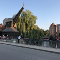 A picture taken from a riverside restaurant of an old fashioned wooden crane. Behind it is a massive weeping willow and several old orange and white bricked buildings with shingled roofs.
