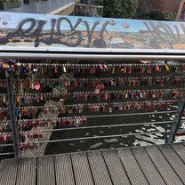A picture of a bridge above a river. There's a panoramic picture of the town of Luneburg that's been graffitied over. On the bridge, there are hundreds of love locks hanging off of suspension wire. 