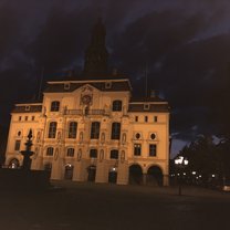 A picture taken at night of an imposing building lit up from below, casting the building in an orange hue. This is the town hall, it has several balconies and a copper roof. There's some marble statues on the facade of the building and the silhouette of a fountain in the foreground. 