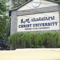 Image of the main sign outside of Christ University that reads "Christ University, deemed to be university," meaning that it is getting accredited. The sign is surrounded by greenery, because campus is very green, allowing for shade and protection from noise.