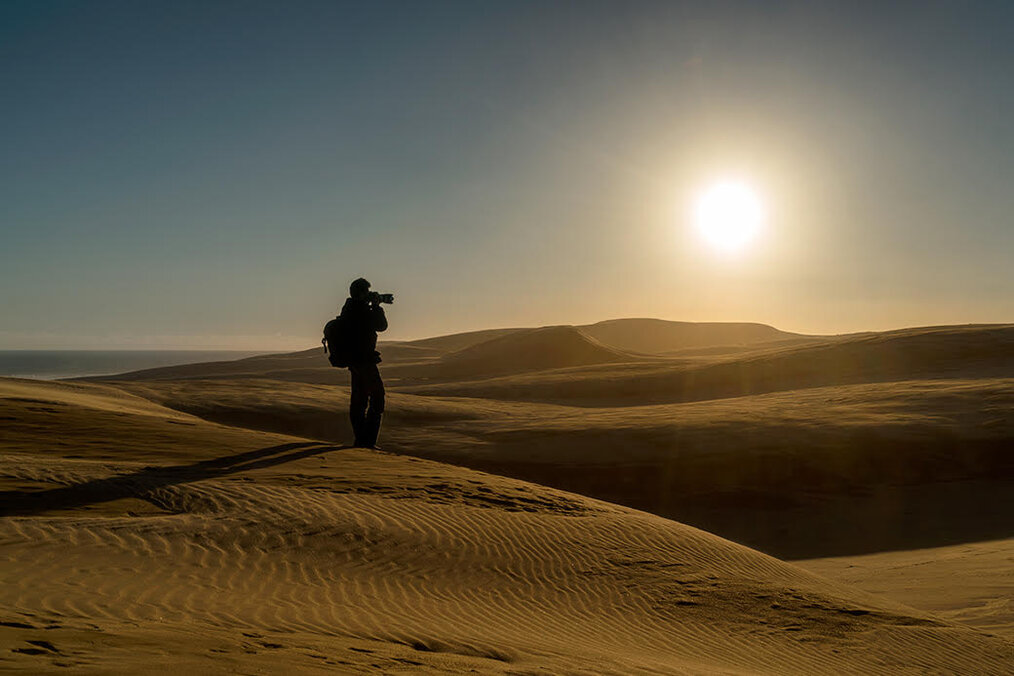 Photo by Colin Murchison A photographer standing on sand dunes with the sun setting behind them