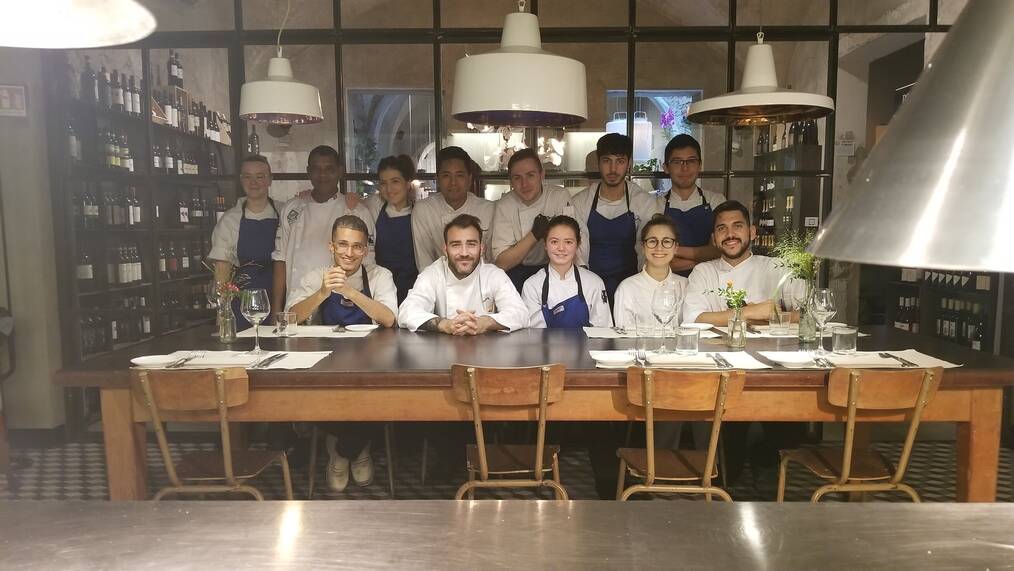 Group of culinary interns wearing chef uniforms and posing around a table.