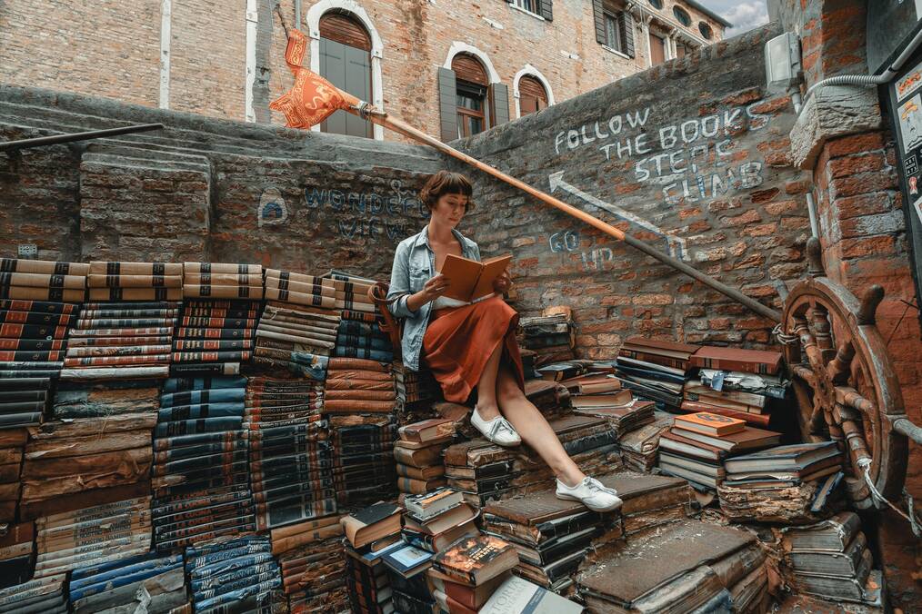 Woman reading a book sitting on a pile of books.