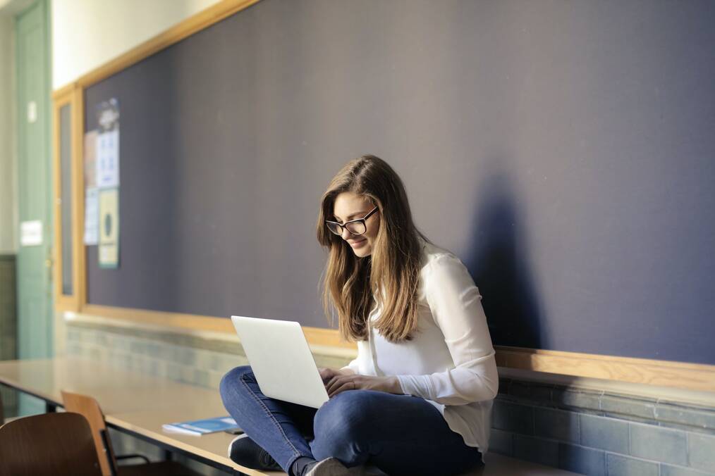 Person sitting on desk in classroom while using laptop