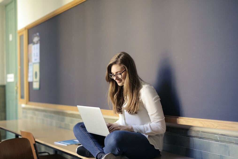 Person sitting on desk in classroom while using laptop