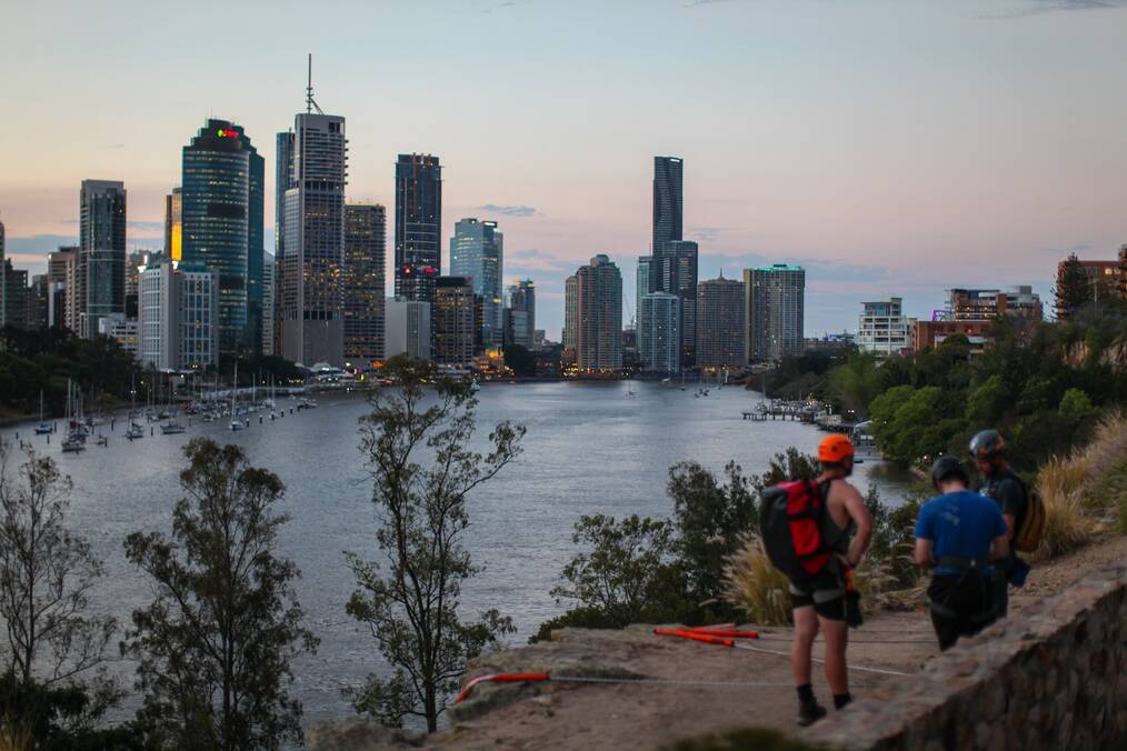 Three men stand beside a river with the cityscape of Brisband, Australia in the background.