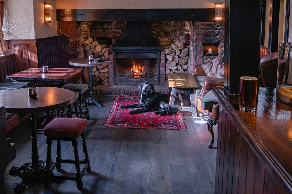 A black dog lays on a rug in front of a fire in an English pub.