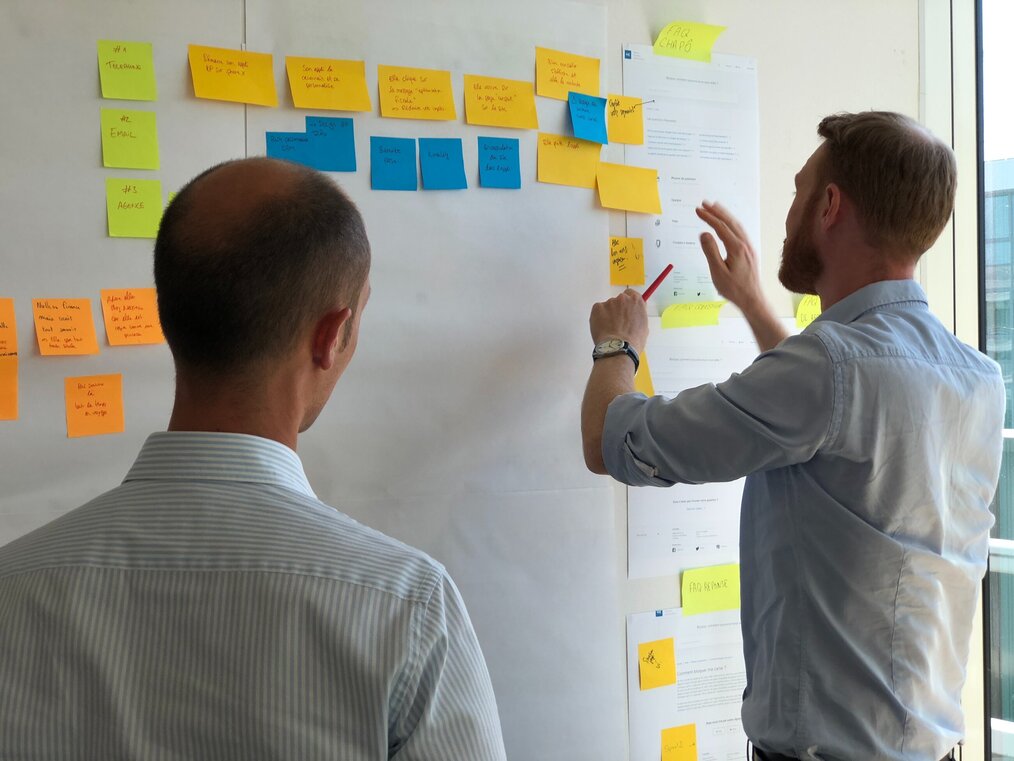 Photo by Bonneval Sebastien on Unsplash Two men look at white board with post-it notes on it.