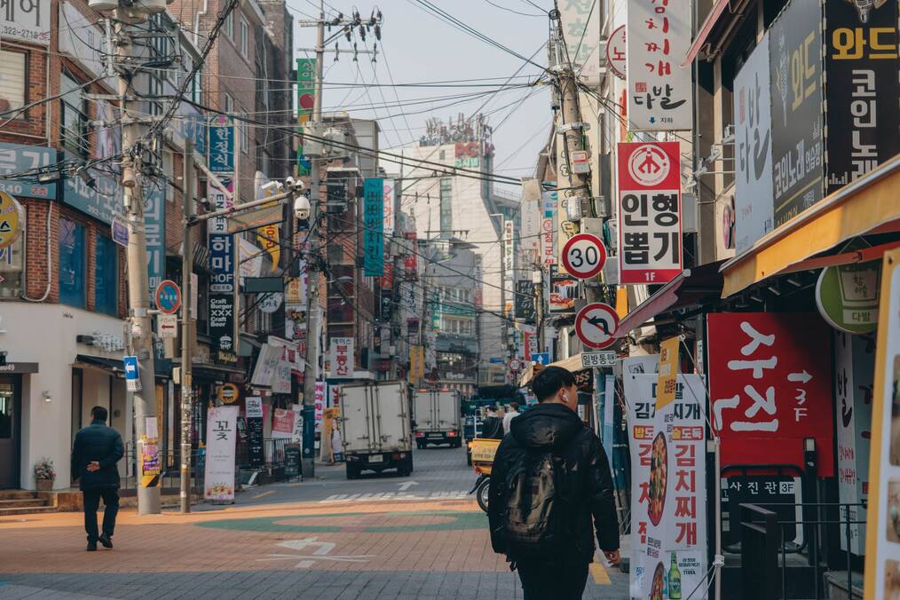 Man walking down Seoul street with many signs and power lines.