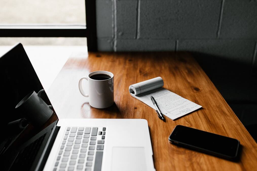 A computer with coffee, a notepad, and an iphone on a desk