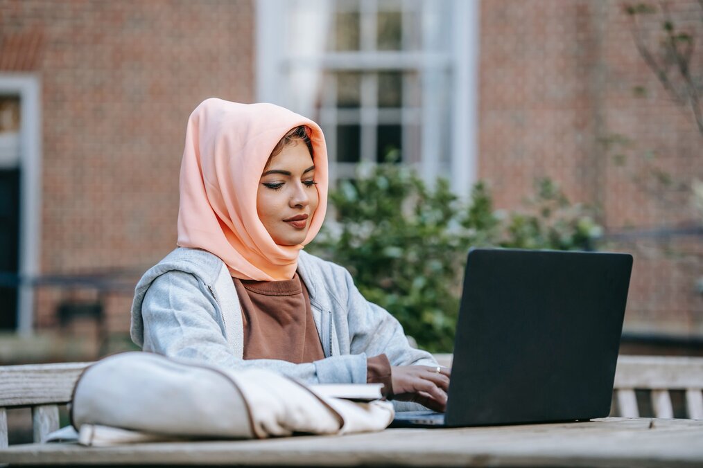 Image curtesy of Keira Burton, Pexels A digital nomad woman with a pink head covering doing virtual assistant job on her laptop