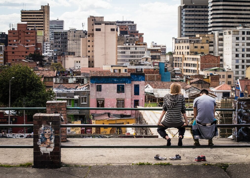 Photo by Delaney Turner on Unsplash Two people sitting on a iron fence with city buildings in the background.
