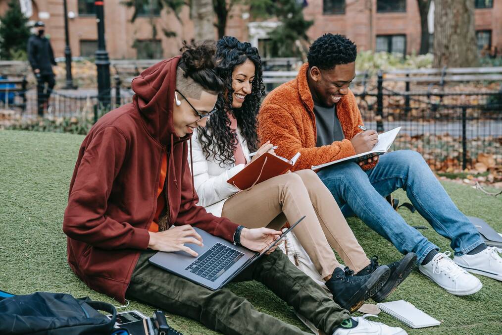 Three people sit on the grass looking at their books and laughing.