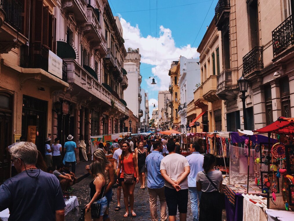 Photo by Rafael Guimarães on Pexels Groups of people stand around at an outside street market in Argentina.