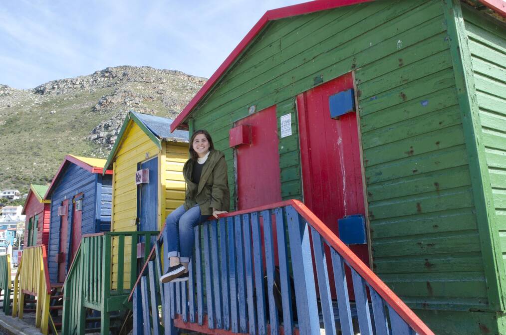 Fernanda sitting on the railing of a colorful hut on Muizenberg beach near Cape Town, South Africa