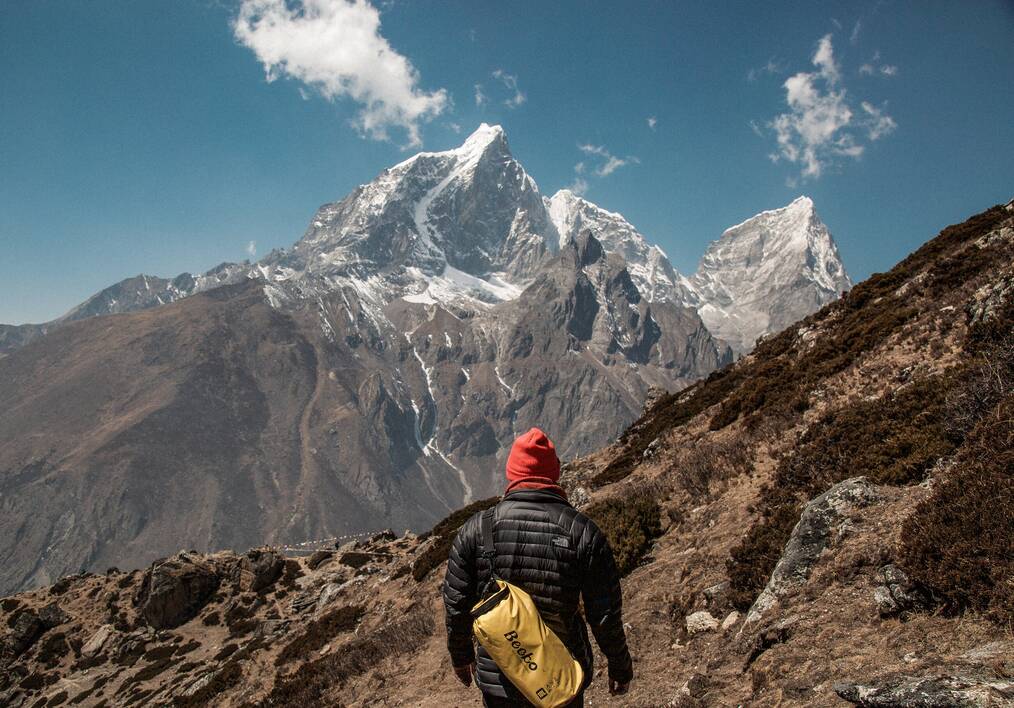 A person wearing a red beanie, a North Face puffer, and a yellow bag faces a mountain summit 