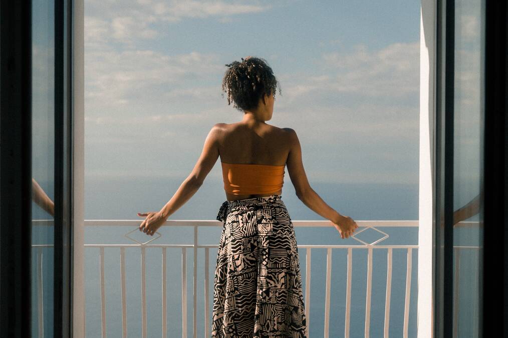 A person in an orange tub top and black and white patterned skirt staring off balcony by the sea
