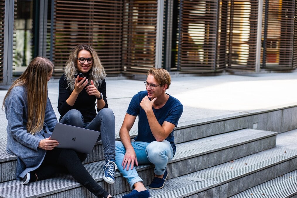 Photo by Buro Millenial on Pexels Three students sit on stairs outside with a laptop.