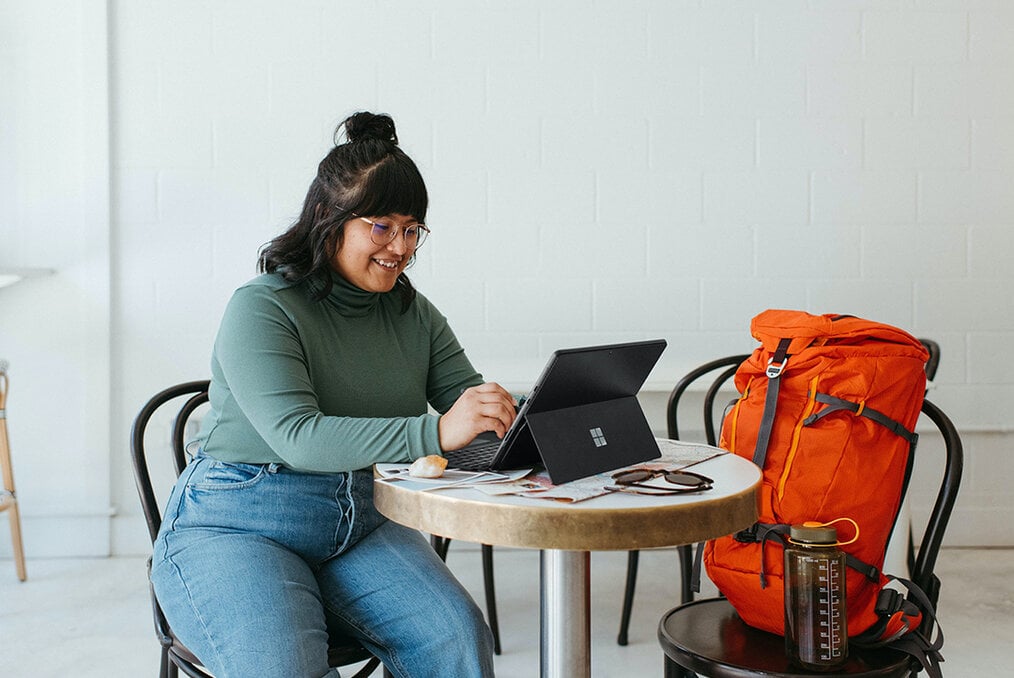 A person sitting at a small round table on a surface tablet with their backpack in the seat next to them