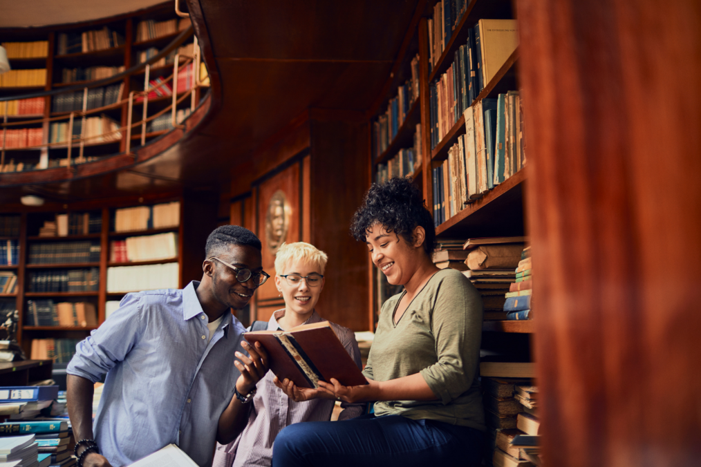 Three people in a library reading a book together smiling and laughing