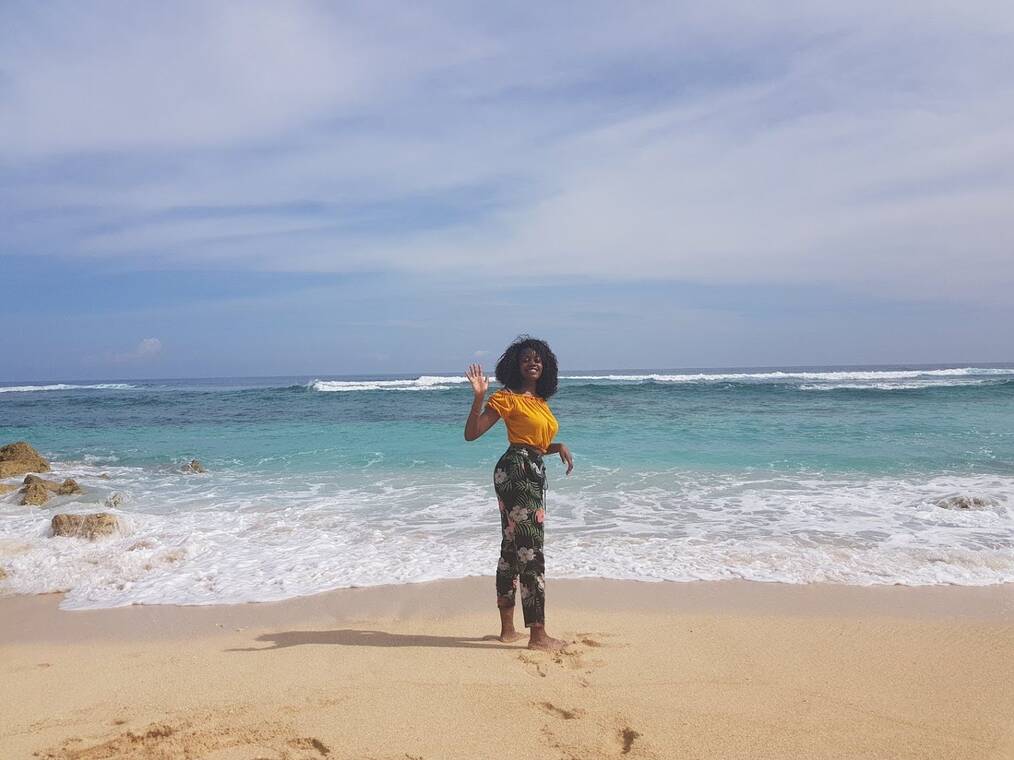 A person in a yellow top and colorful pants waving and smiling on the beach in Bali