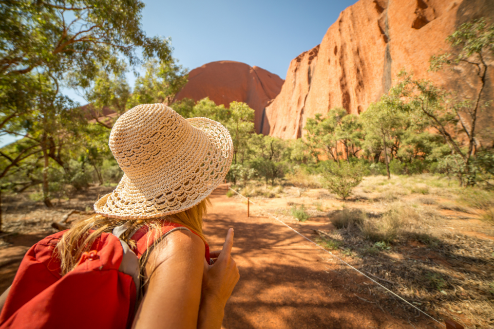 A person with a red backpack, blonde hair and a crochet hat looks at the Australian bush