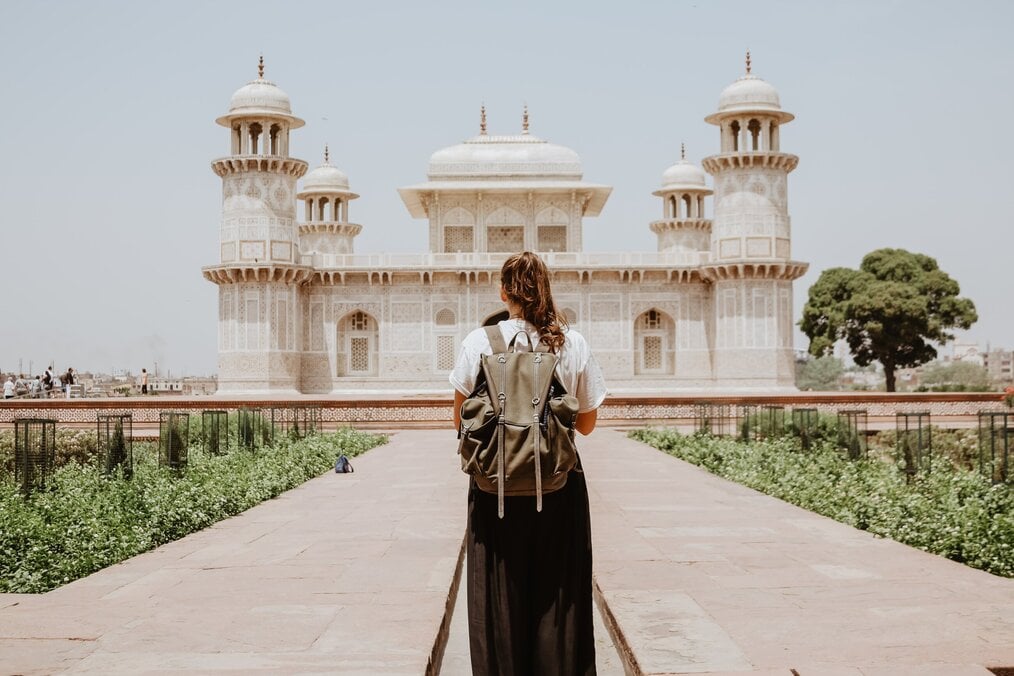 A woman wearing a backpack stands with her back to the camera in front of a tomb in India.