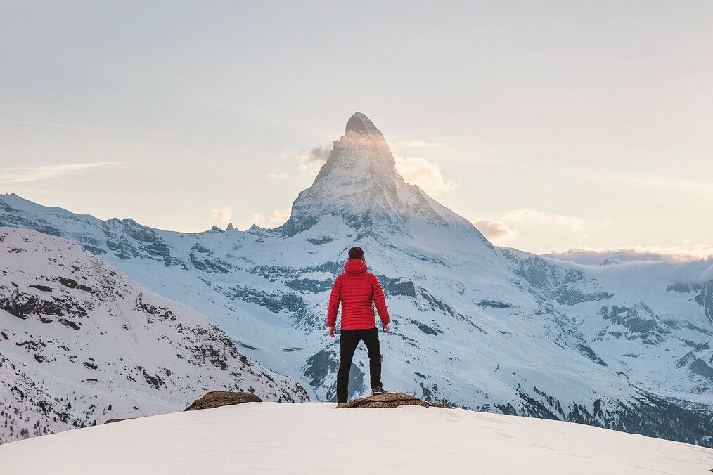 Image curtesy of Joshua Earle, Unsplash A person wearing a red puffer jacket standing on a summit staring at The Matterhorn mountain in the alps