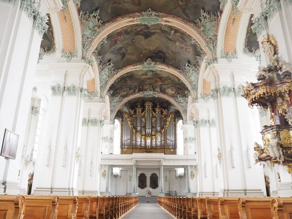 The inside of the St. Gallen Cathedral with white pillars and blue intricate detailing to a heavenly mural on the ceilings