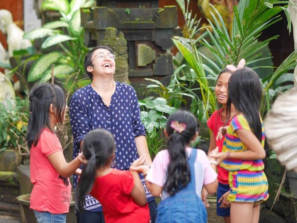Ghada H., Volunteer Programs Bali A smiling volunteer stands holding hands with a group of children.