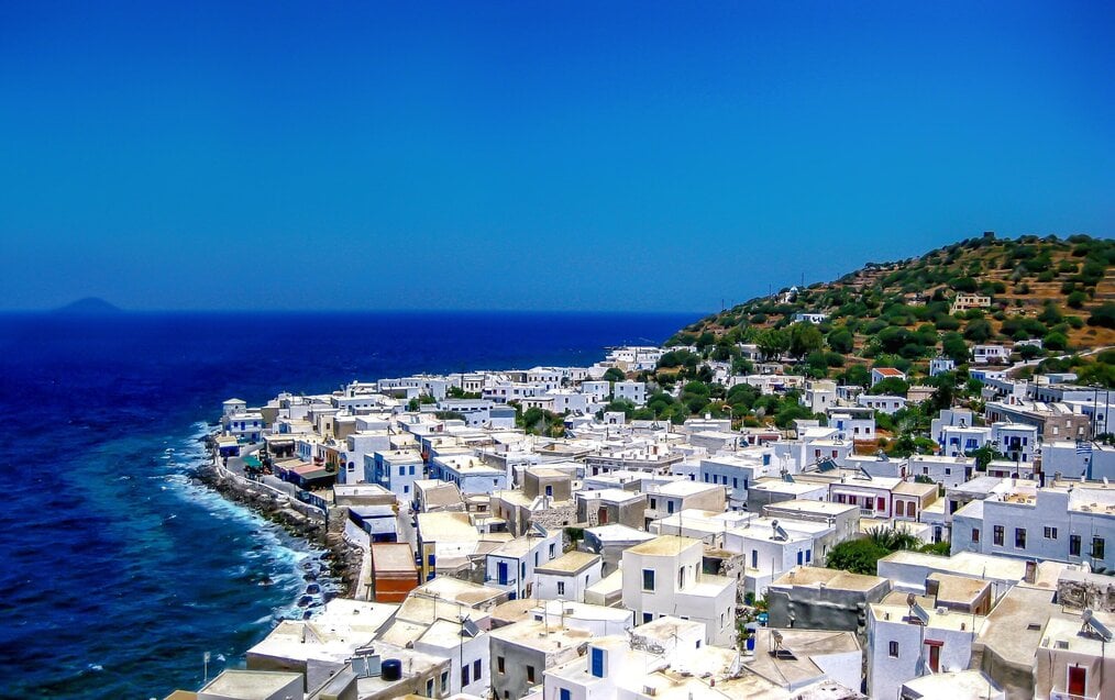 Photo by Nicolas Lysandrou on Unsplash White houses sit on a hill above blue water in Greece.