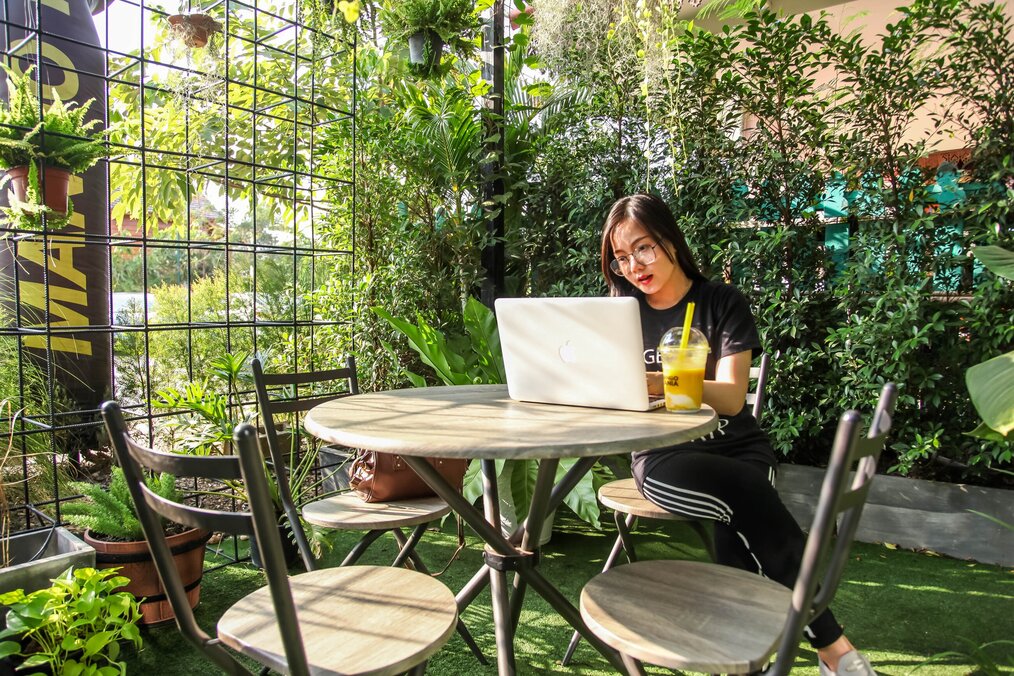 Photo by DoDo PHANTHAMALY A woman sits at a table with her laptop surrounded by green plants.