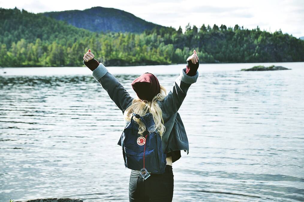 A woman stands with her back to the camera, raising her arms in the air. There is a lake and forest in the background.