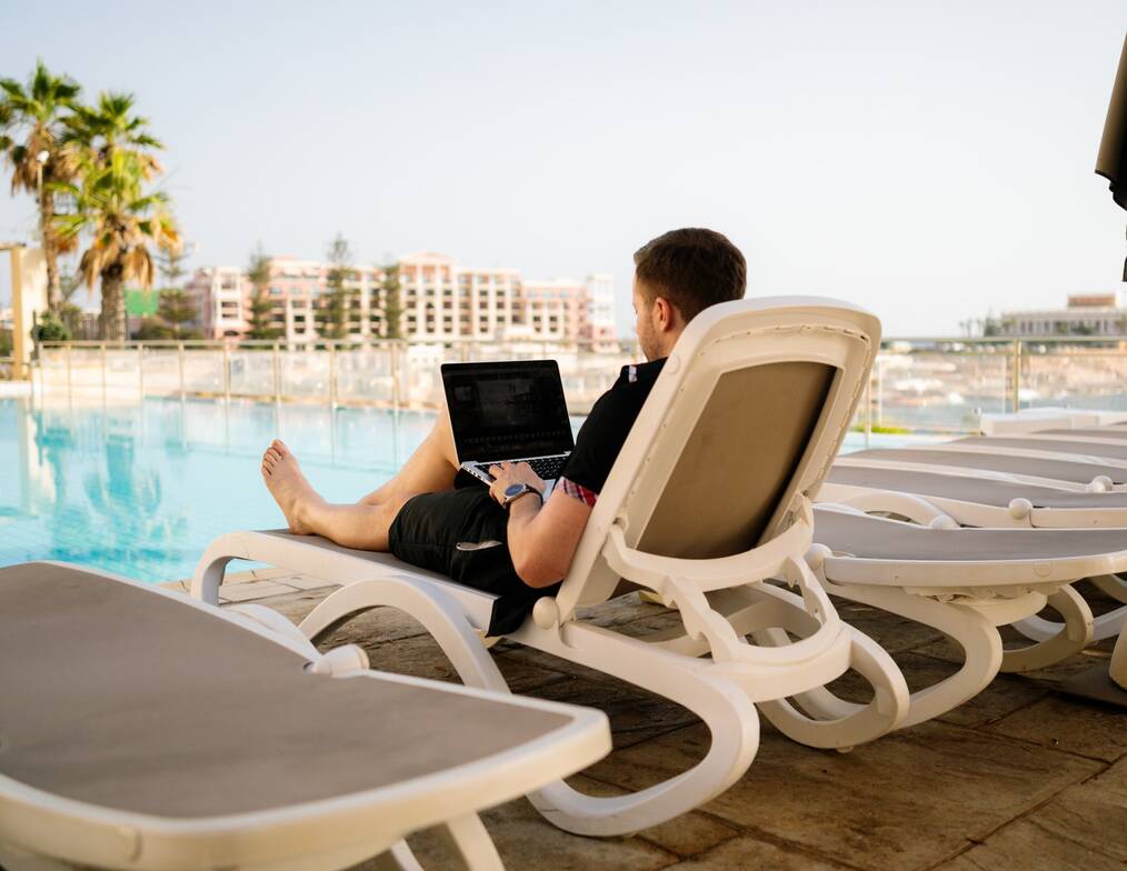 A man sits on a deck chair with a laptop on his lap, a pool is in the background.