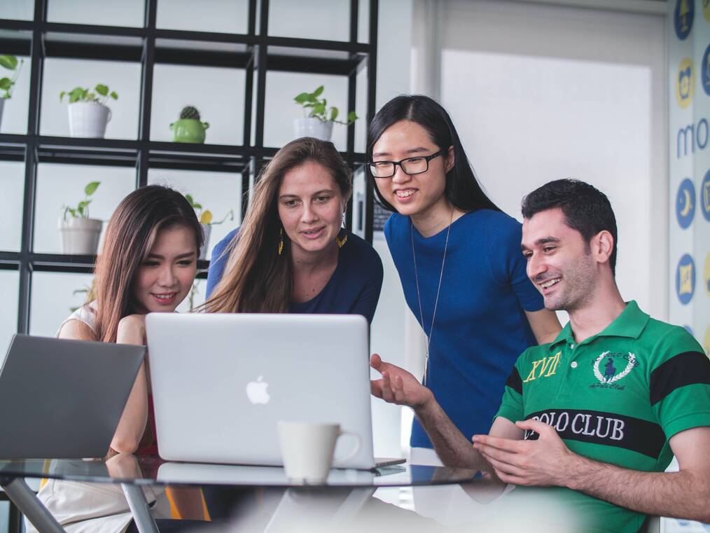 A group of three women and a man stand around a laptop looking at it and smiling.