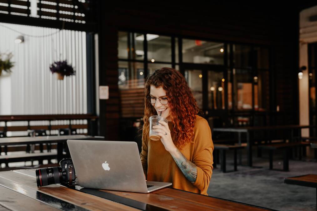 A smiling woman sips a drink through a straw as she works on her laptop.