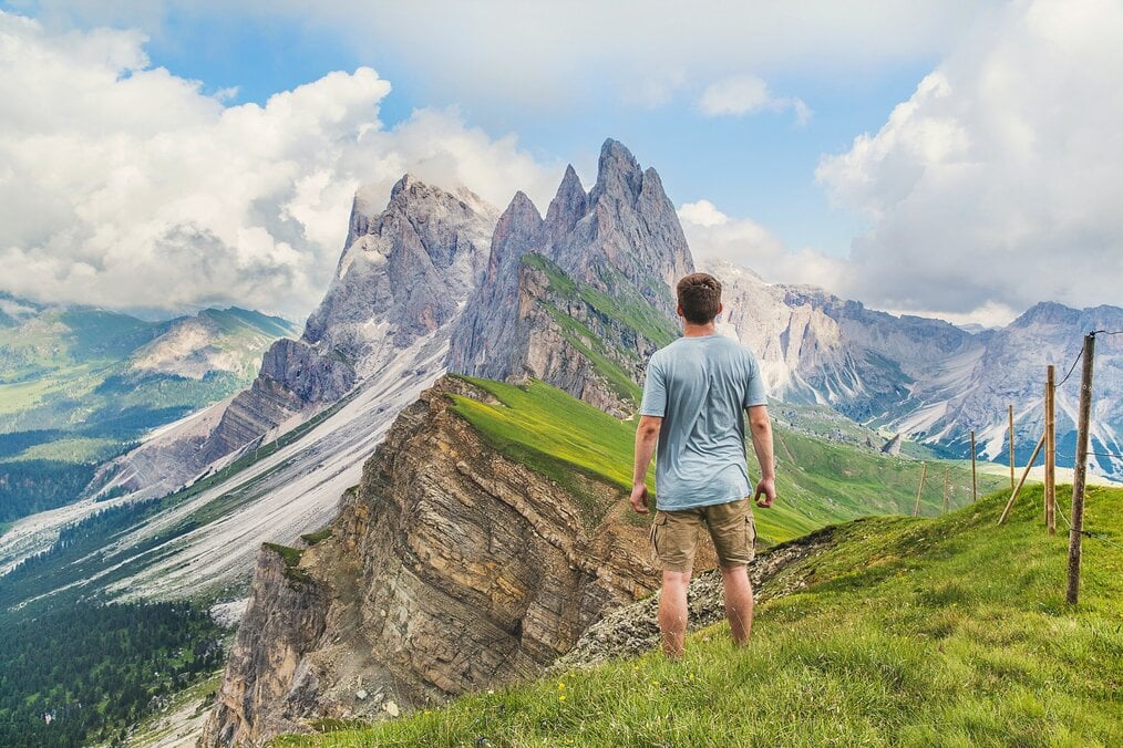 Man stands on green hill with back to camera while looking at large jagged mountains.