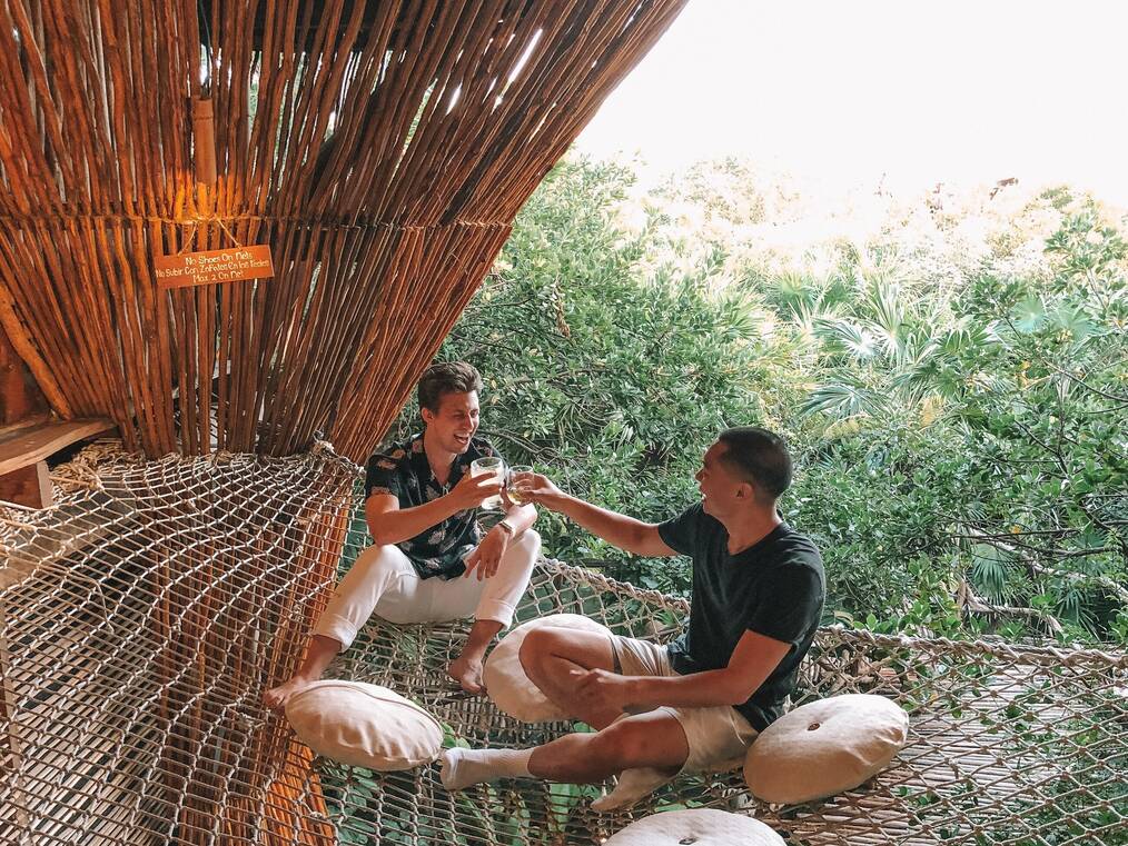 Two men sitting on rope netting cheers their glasses and laugh.