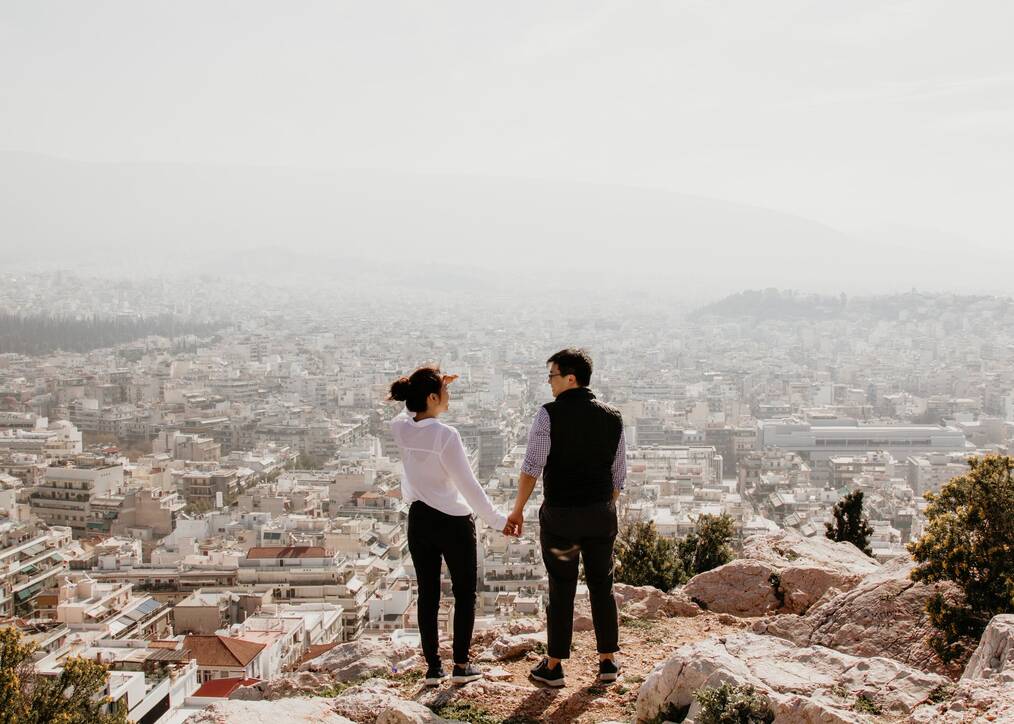 A couple holds hands standing on a rock overlooking the city of Athens, Greece.