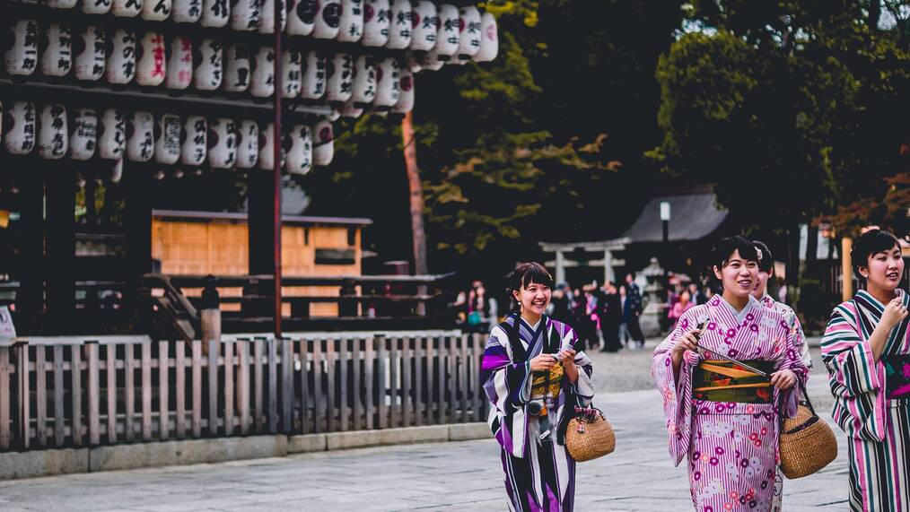 Women dressed in traditional Japanese clothing stand and smile outside.