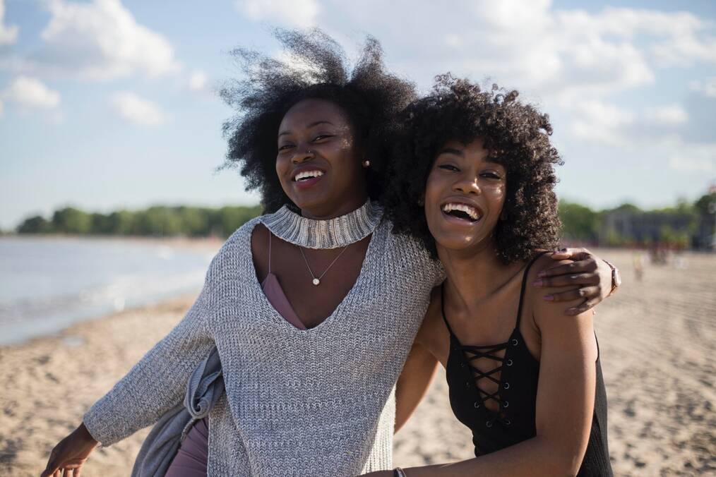 Two women with their arms around each other smile on a beach.