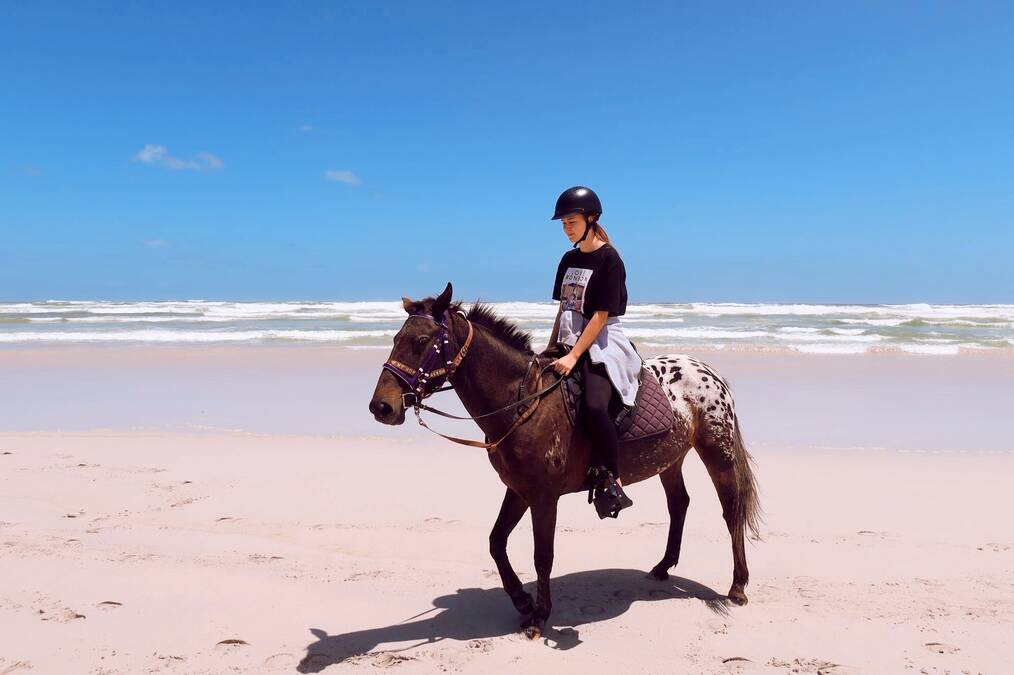A woman rides a brown horse on a white sand beach.