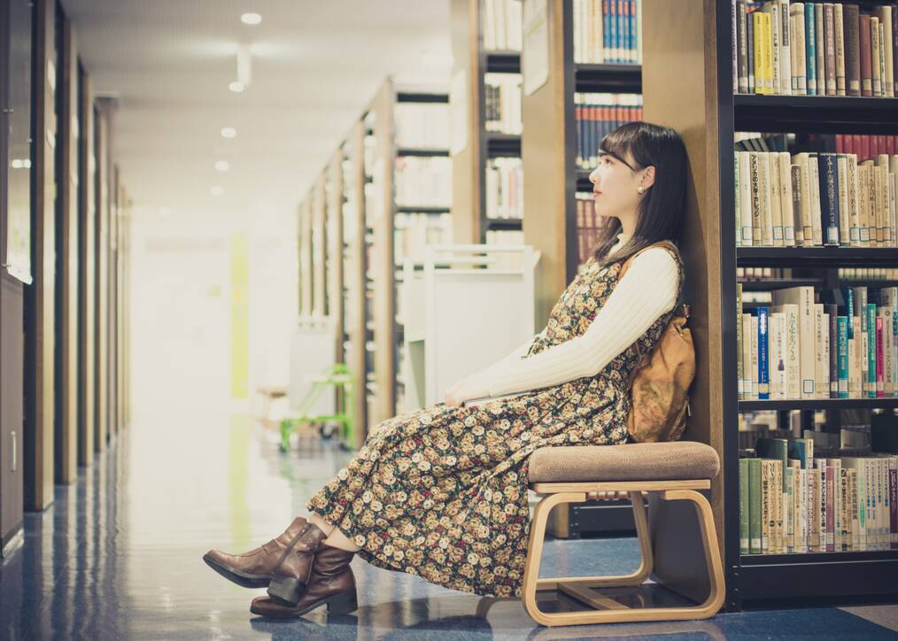 A woman wearing a backpack sits on a chair in a library.
